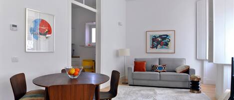 Een moderne woonkamer met een grijze bank, een ronde houten eettafel met stoelen, en twee kunstwerken in lijsten aan de muur.