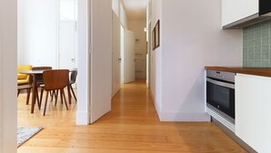 A narrow hallway with wooden flooring leading to a dining area with a yellow sofa and wooden chairs. A kitchen with a stove and cabinets is visible on the right. - Alfama Deluxe by Homing (Lisbon)