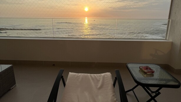 Un balcon offrant une vue sur le coucher de soleil sur la mer, un fauteuil confortable et une petite table agrémentée d’un objet décoratif.