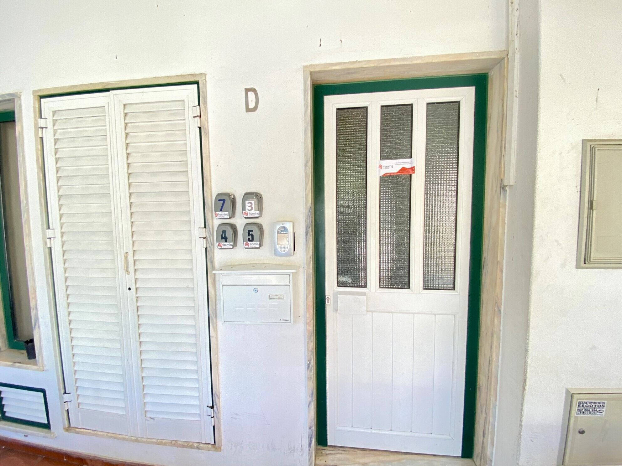 A white door with a green frame, a window with a metal grill, and a set of four house numbers (7, 3, 4, 5) above the door.