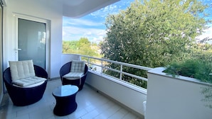 Un balcone con due poltrone in vimini, un tavolino rotondo e una porta vetrata, con vista su alberi e edifici.