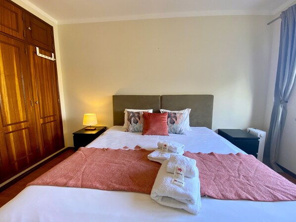 A hotel room with a bed, a nightstand with a lamp, a wooden wardrobe, and a window with curtains. - Vilamoura Fresh With Pool by Homing (Loulé)