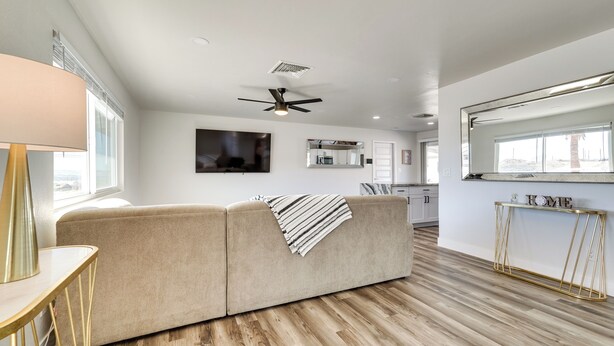 A modern living room with a beige sofa, a wooden floor, a ceiling fan, and a kitchen area in the background. - Patio + Private Casita: Lake Havasu City Home! (Lake Havasu City)