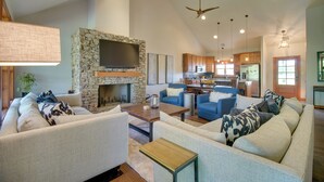 A spacious living room with a stone fireplace, a large flat-screen TV, and a dining area with wooden cabinets and a kitchen in the background.