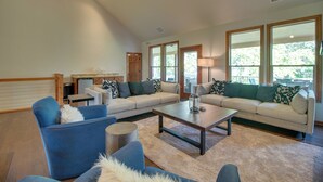 A spacious living room with a large sofa, a coffee table, and a blue armchair. There are large windows providing natural light and a view of greenery outside.