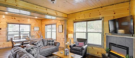Interior acogedor de una cabaña de troncos con chimenea, un televisor montado encima, comedor con mesa y sillas, y zona de estar cómoda con sofá y sillones.