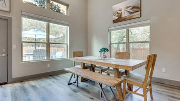 A dining area with a wooden table, benches, and chairs. There are large windows providing natural light and a view of the outdoors.