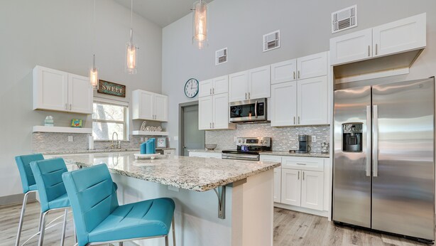 A modern kitchen with white cabinets, a stainless steel refrigerator, a large island with bar stools, and pendant lighting. - Modern Canyon Lake Vacation Home w/ Furnished Deck (Canyon Lake)