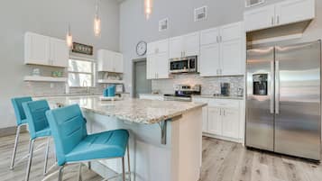 A modern kitchen with white cabinets, a stainless steel refrigerator, a large island with bar stools, and pendant lighting.