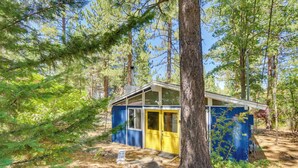 A small cabin with a blue exterior and yellow door, surrounded by trees and a wooden deck. - Pet-friendly Big Bear Cabin, Near Hiking & Skiing! (Big Bear City)
