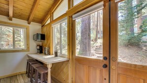 A wooden interior with a kitchen counter, stools, and a door leading to an outdoor area with trees. - Pet-friendly Big Bear Cabin, Near Hiking & Skiing! (Big Bear City)