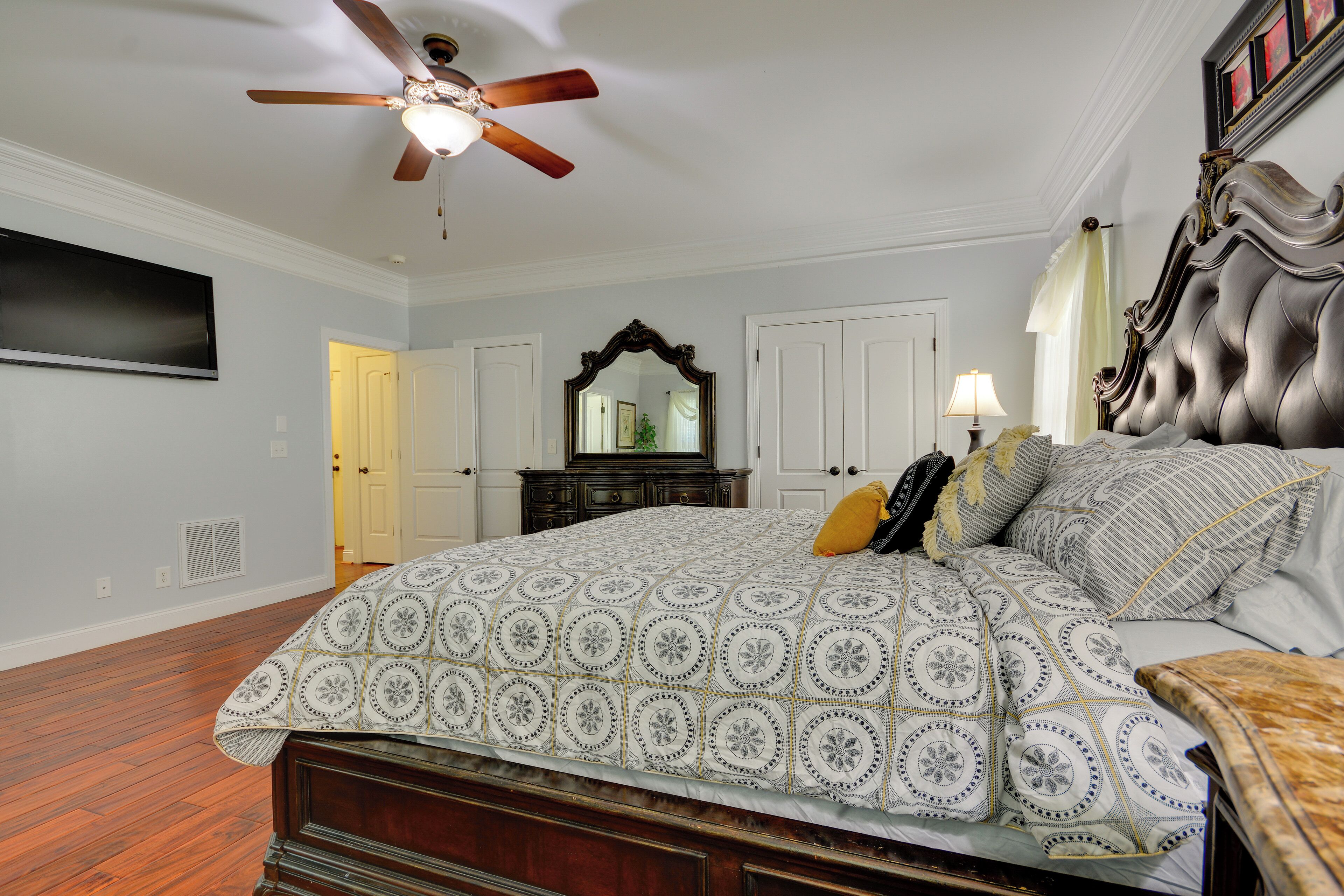 A bedroom with a large bed, a ceiling fan, a flat-screen TV, a dresser, and a mirror.
