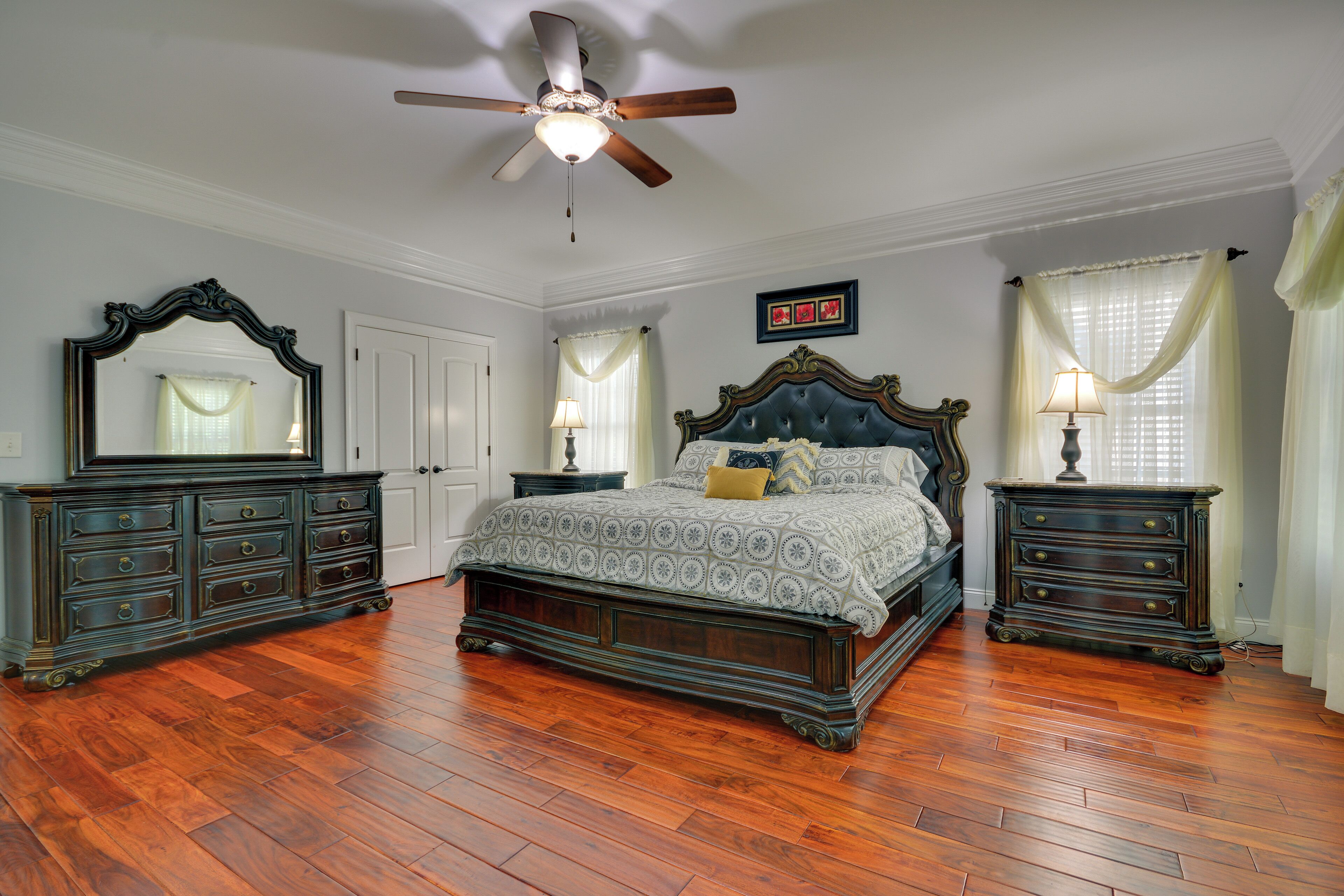 A bedroom with a large bed, two nightstands, a dresser, a ceiling fan, and a window with curtains.