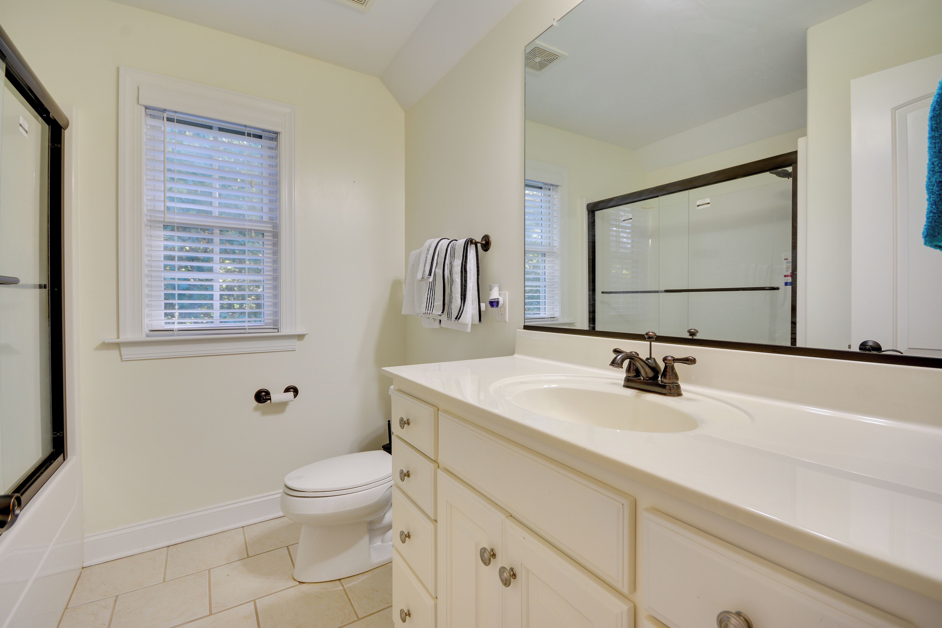 Une salle de bain avec des toilettes blanches, un grand lavabo avec un miroir et une fenêtre avec des stores.