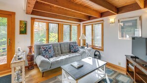 A cozy living room with a grey sofa, a coffee table, and a television. There are large windows allowing natural light and views of the outdoors. - Ellsworth Home ~ 18 Mi to Acadia Nat'l Park! (Ellsworth)
