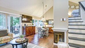 A spacious living area with a sofa, coffee table, and dining table. A kitchen with wooden cabinets and a fireplace is visible in the background. - Gilford Townhome w/ Lakefront Beach & Pool Access! (Gilford)