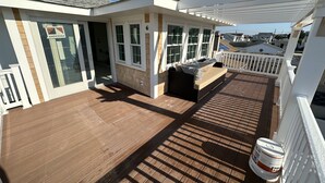 Una terraza techada al aire libre con piso de madera, un área de descanso con cojín y barandal blanco.