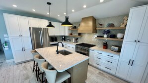 Una cocina moderna con armarios blancos, una isla central con taburetes de bar, un refrigerador de acero inoxidable, una estufa a gas e iluminación colgante.