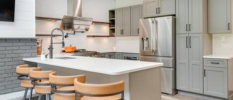 A modern kitchen with a central island, bar stools, stainless steel appliances, and light grey cabinetry.