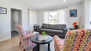 A modern living room with a grey sofa, colorful patterned chairs, a round black table with decorative items, and a view of a house through large windows. - Dom Rodzinny Lakeview Getaway - Walk to Lake Huron (Rogers City)