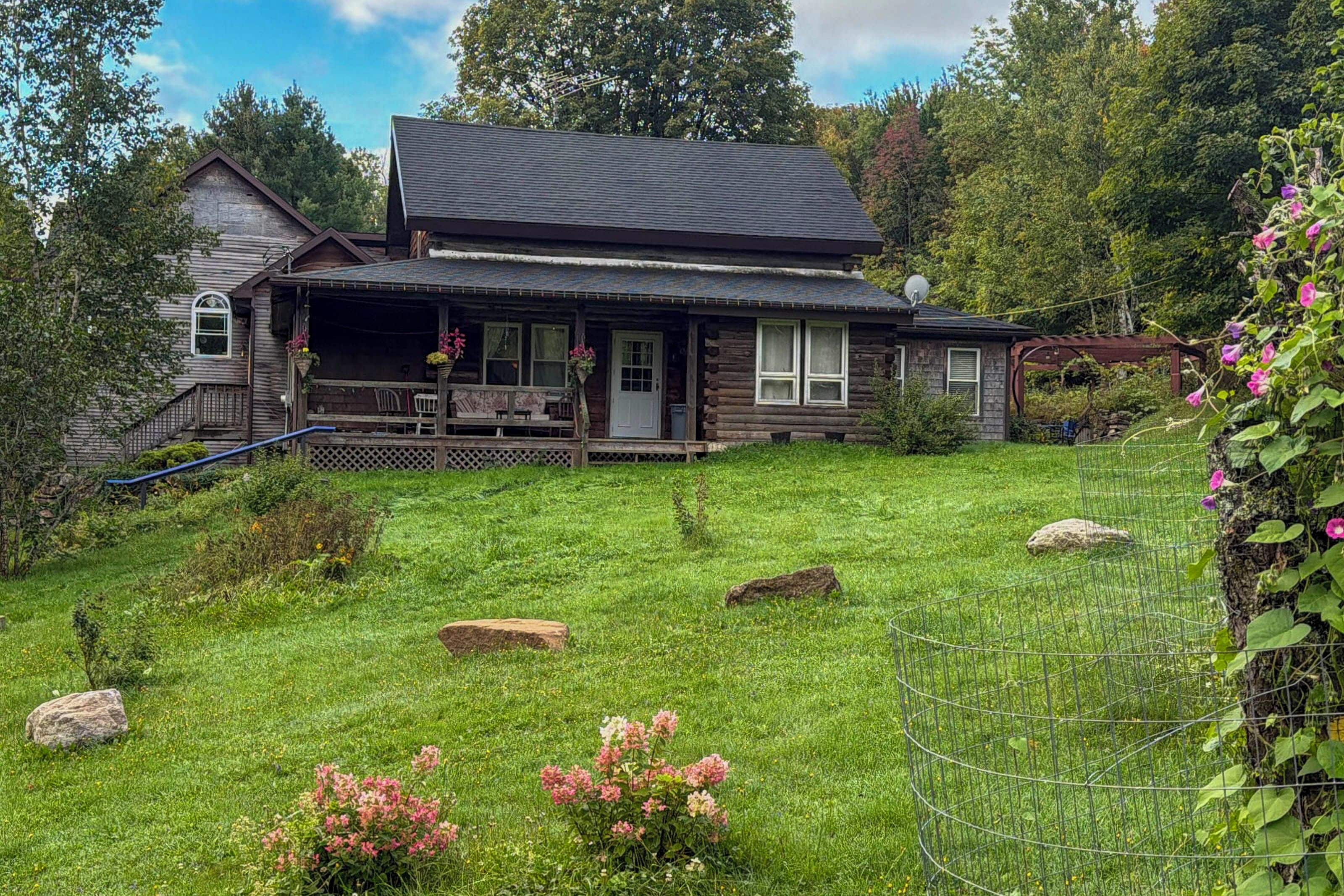 A two-story log cabin with a porch, surrounded by a grassy yard with rocks and flowers.
