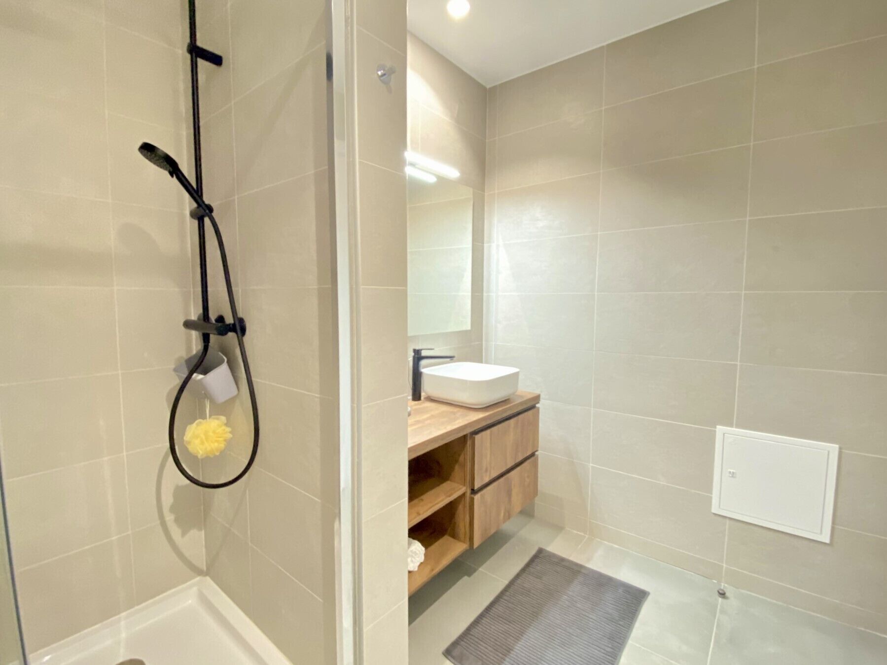 A bathroom with beige tiles, a white sink, wooden cabinets, a shower with a handheld showerhead, and a mirror.