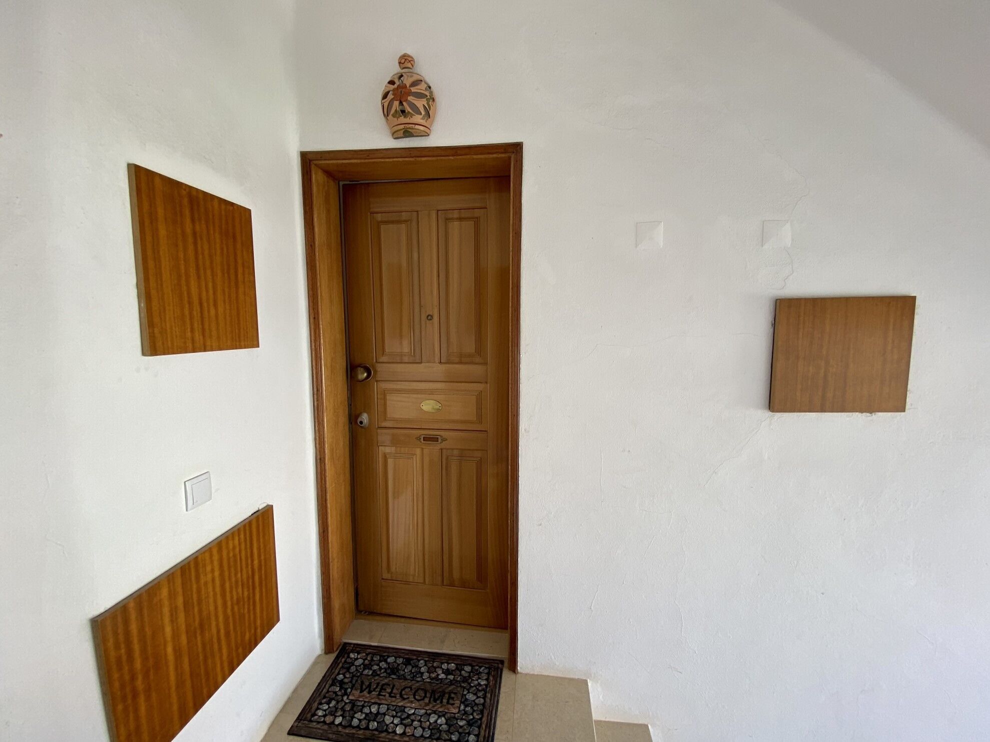 Une porte en bois avec un paillasson, un petit objet décoratif placé au-dessus de la porte et un panneau en bois sur le mur.