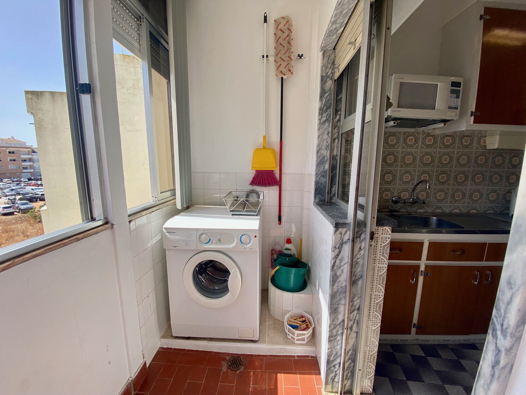 Un balcón pequeño con lavadora, escoba, mopa y microondas en la zona de la cocina contigua.