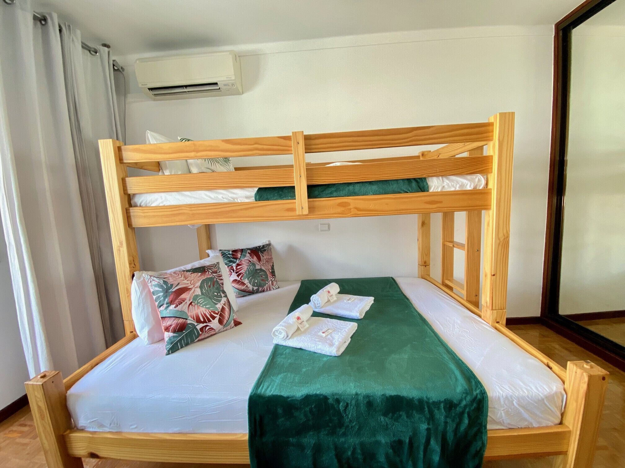 A bunk bed with a green blanket and tropical pillows, an air conditioning unit, and a window with wooden frames.