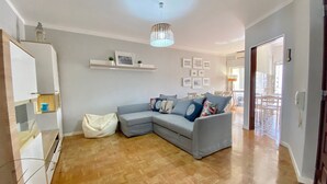 A modern living room with a grey sectional sofa, wooden flooring, a white wall with shelves, and a hanging light fixture. - Quarteira Beach Apartment by Homing (Loulé)