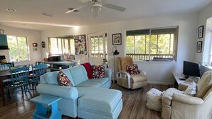 A spacious living room with a light blue sectional sofa, a beige armchair, and a blue coffee table. There is a dining area with a table and chairs, and a kitchen area with a stove and cabinets. - Peaceful location! Close to everything! Bring the family and dogs! Snowbirds! (Bon Secour)