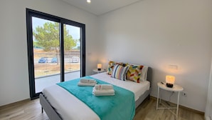 Une chambre d’hôtel moderne avec un grand lit, une vue dégagée sur l’extérieur grâce à une porte coulissante, une petite table de chevet avec une lampe et une fenêtre donnant sur le paysage.