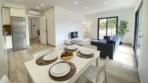 Une cuisine moderne dotée d’un réfrigérateur encastré, un coin repas avec une table blanche dressée pour quatre personnes, et un salon avec un canapé et un téléviseur.