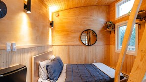Una habitación acogedora tipo cabaña de madera, con una cama, un espejo redondo y una ventana.