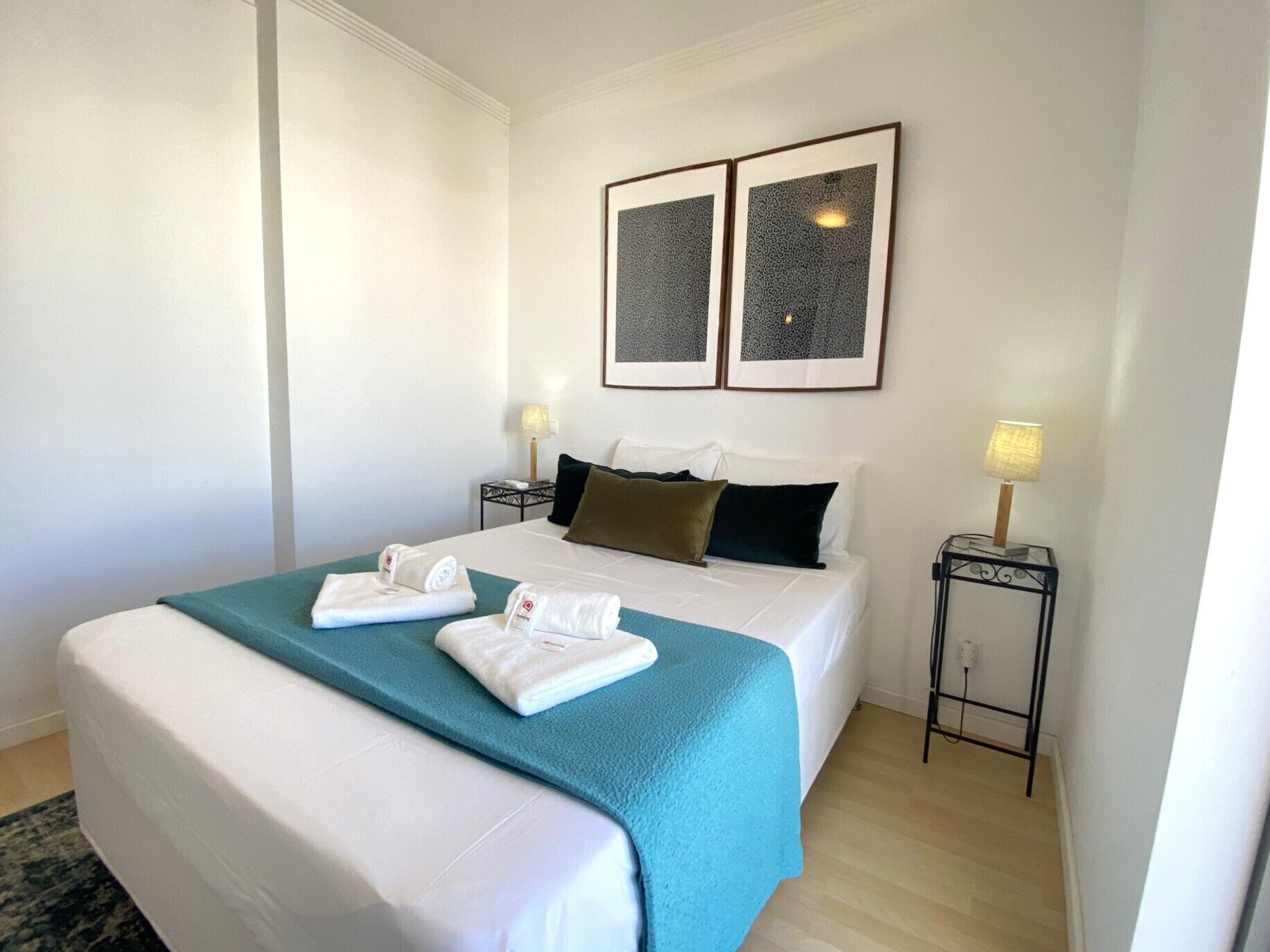 A hotel room with a bed, two bedside tables, a nightstand, a lamp, and two framed pictures on the wall.