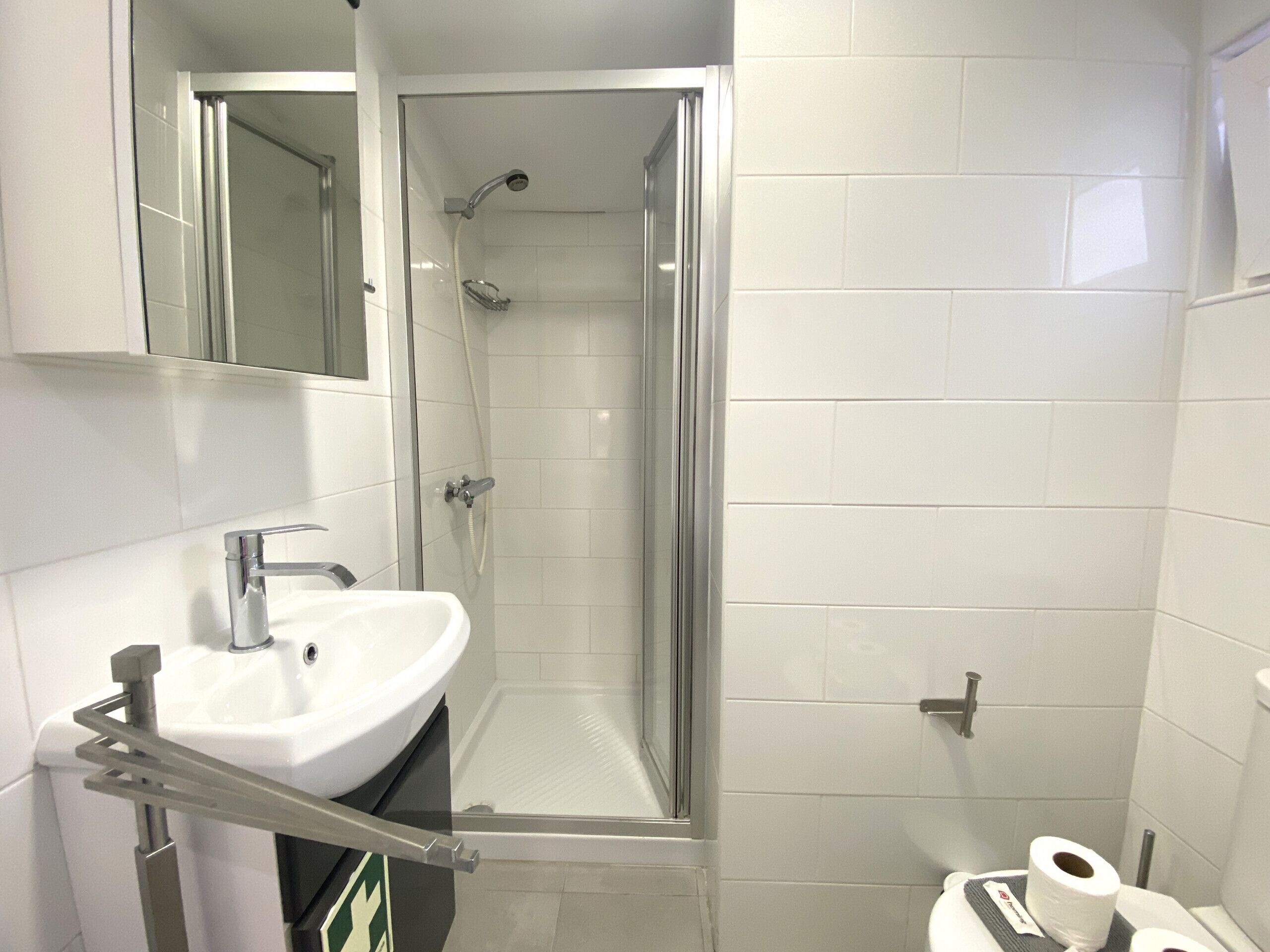 Un baño con ducha de vidrio, lavamanos con toallero y retrete con papel higiénico.