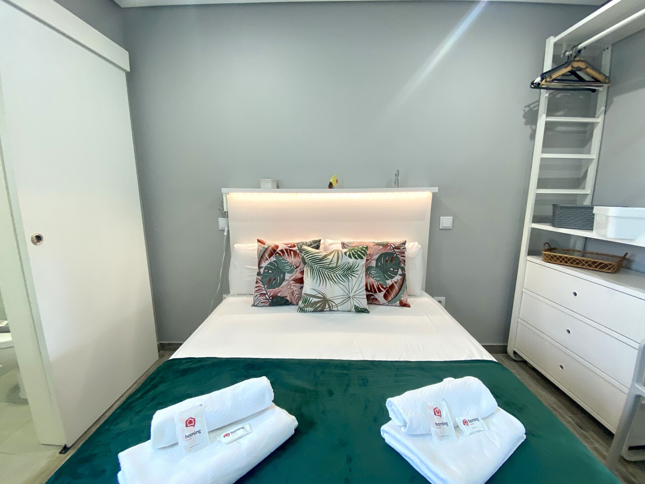 A neatly made bed with a green bedspread, two white towels with 'homing' branding, and tropical patterned pillows. A white dresser with a basket and a white coat rack with a hanger are visible.