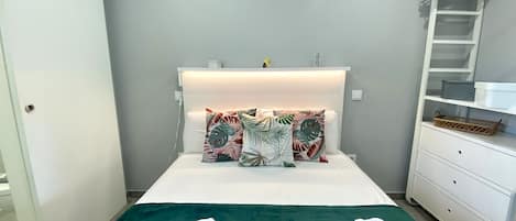 A neatly made bed with a green bedspread, two white towels with 'homing' branding, and tropical patterned pillows. A white dresser with a basket and a white coat rack with a hanger are visible.