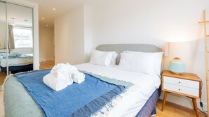 Ein ordentlich bezogenes Bett mit einer blau-weißen Tagesdecke, ein Nachttisch mit Lampe und ein großer Spiegel an der Wand.