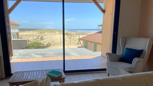 Une pièce dotée d’une grande fenêtre donnant sur une plage et l’océan. On y trouve un fauteuil blanc avec un coussin bleu et une petite table en bois sur laquelle est posé un livre.