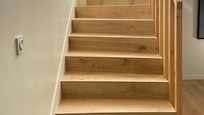 Un escalier en bois au design moderne, menant à un palier où se trouve un interrupteur.