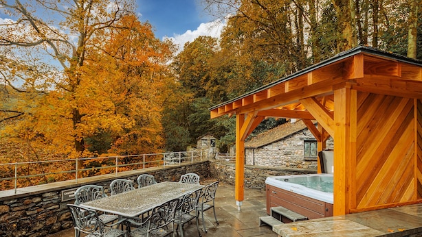 An outdoor patio with stone walls, a wooden pergola, a hot tub, and metal chairs.