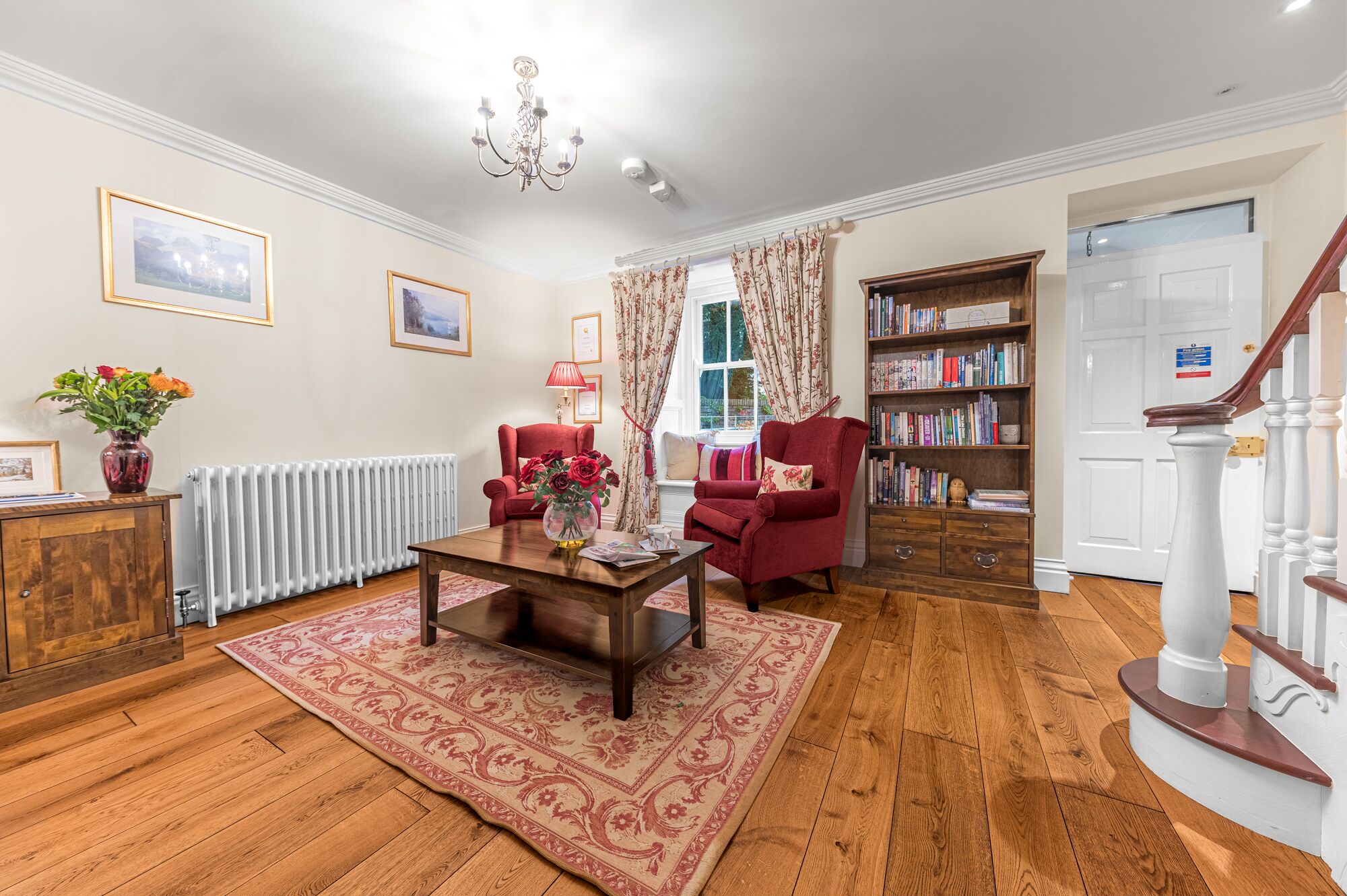 A living room with a wooden floor, a red armchair, a coffee table, a bookshelf, and a staircase.