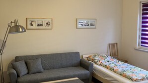 Et lille, godt oplyst rum med en grå sofa, et træbord, en enkeltseng med blomstret dyne og to billeder i rammer på væggen.