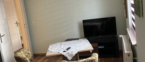 Una habitación pequeña con piso de madera, una mesa con control remoto, dos sillones de estampado floral, una televisión y un piano.