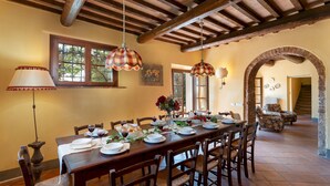 Un comedor con una larga mesa de madera dispuesta para una comida, rodeada de sillas, un techo rústico y una ventana que deja entrar luz natural.