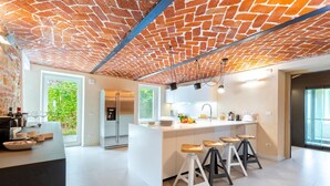 Une cuisine moderne avec un plafond en briques, des meubles blancs, un îlot central doté de tabourets et une vue sur la verdure à l’extérieur.