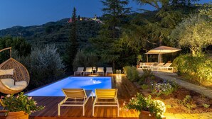 Un espace piscine doté de chaises longues, d’une chaise suspendue et d’un coin salon abrité, le tout entouré d’une végétation luxuriante et offrant une vue sur une colline où se trouve un bâtiment.