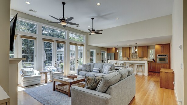 A spacious living area with a large sofa, a coffee table, and a television. There are two ceiling fans, a kitchen with wooden cabinets, and large windows allowing natural light.