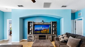 A living room with a large flat-screen TV mounted on a wooden entertainment unit, a brown sofa, and a matching ottoman. - Aster Cove (Kissimmee)